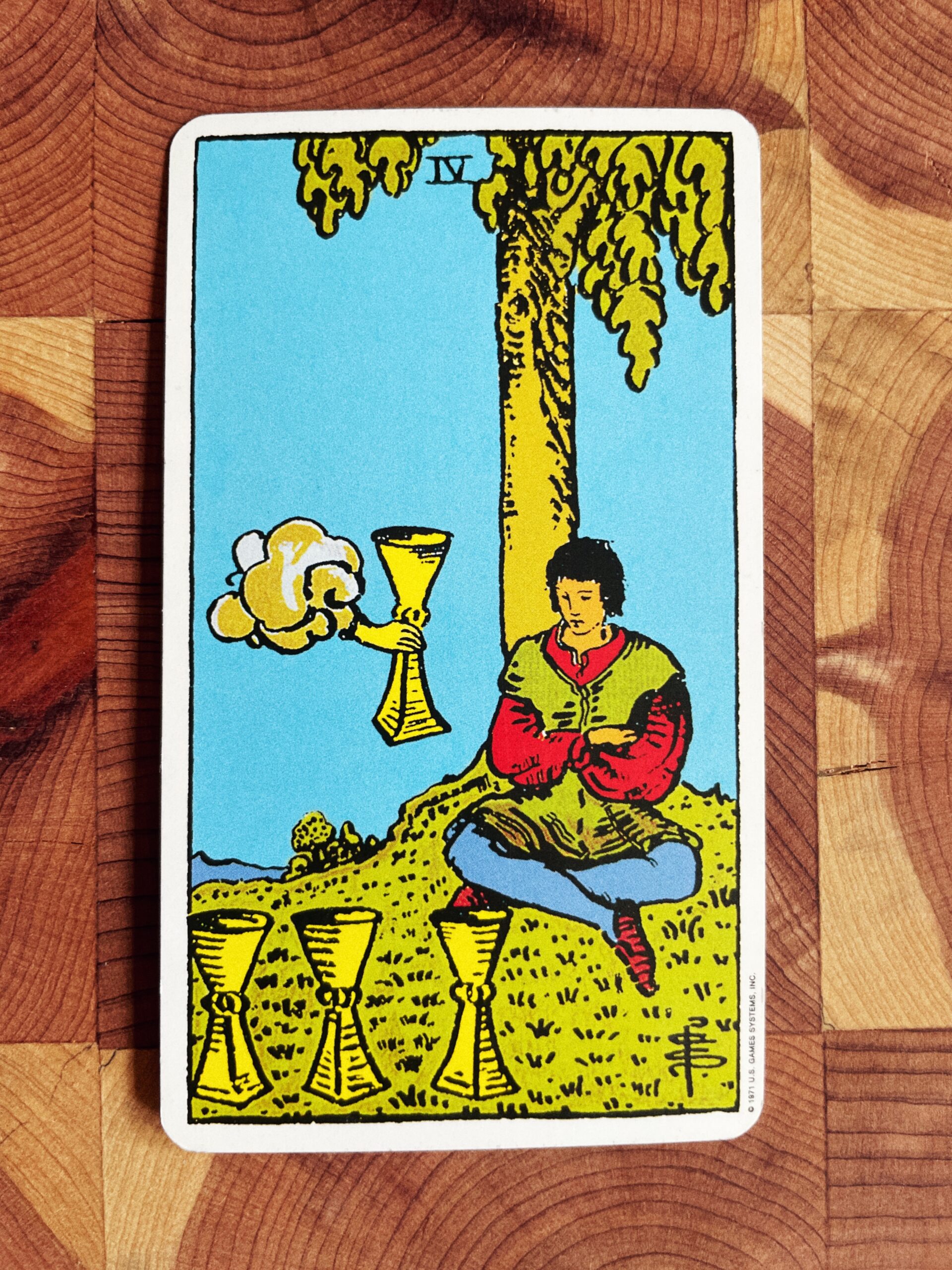 4 of Cups Rider Waite Tarot Deck. A hand outstretches a forth cup to a single man, three cups sit in the foreground. 